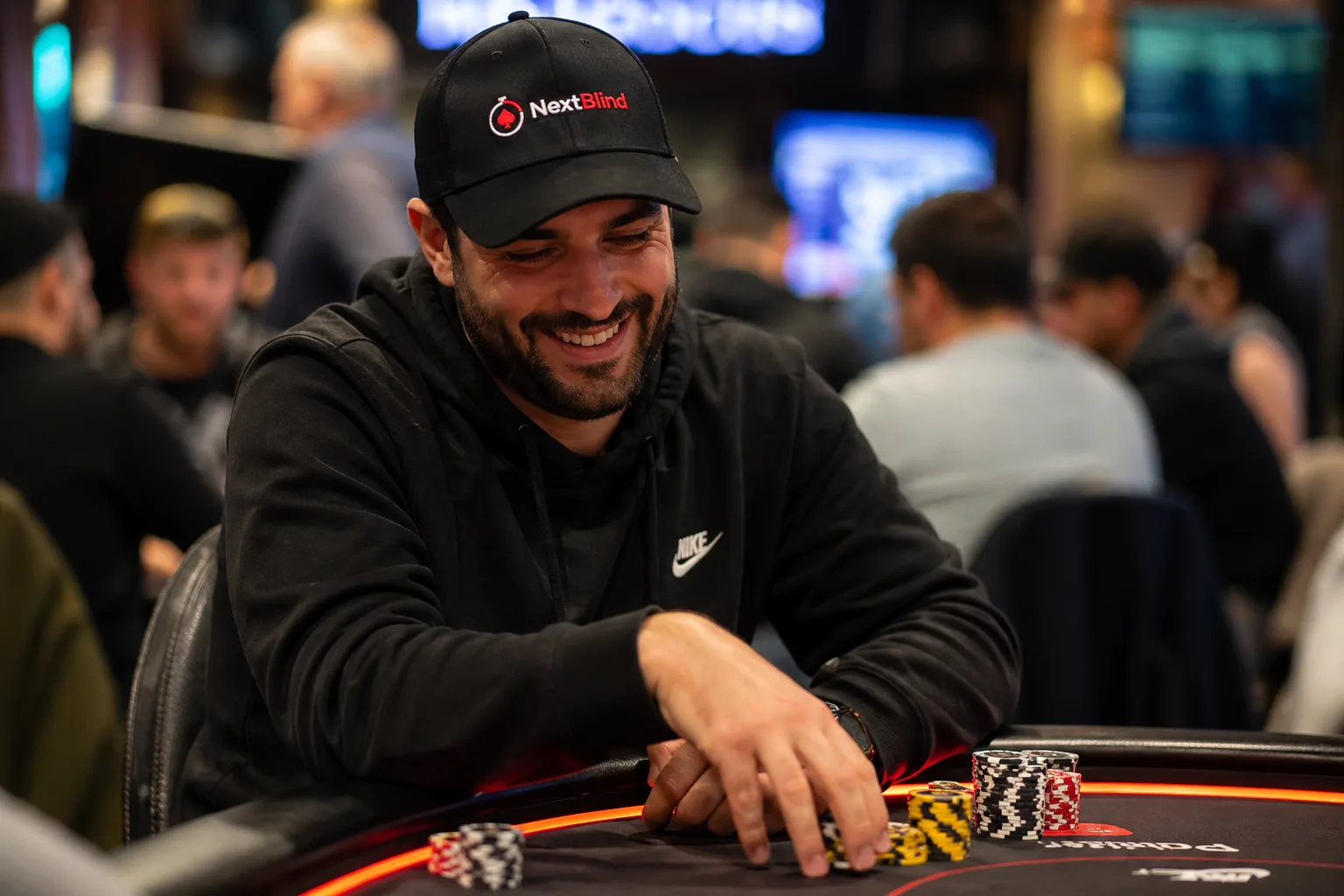 A smiling poker player wearing a NextBlind cap at a live tournament table